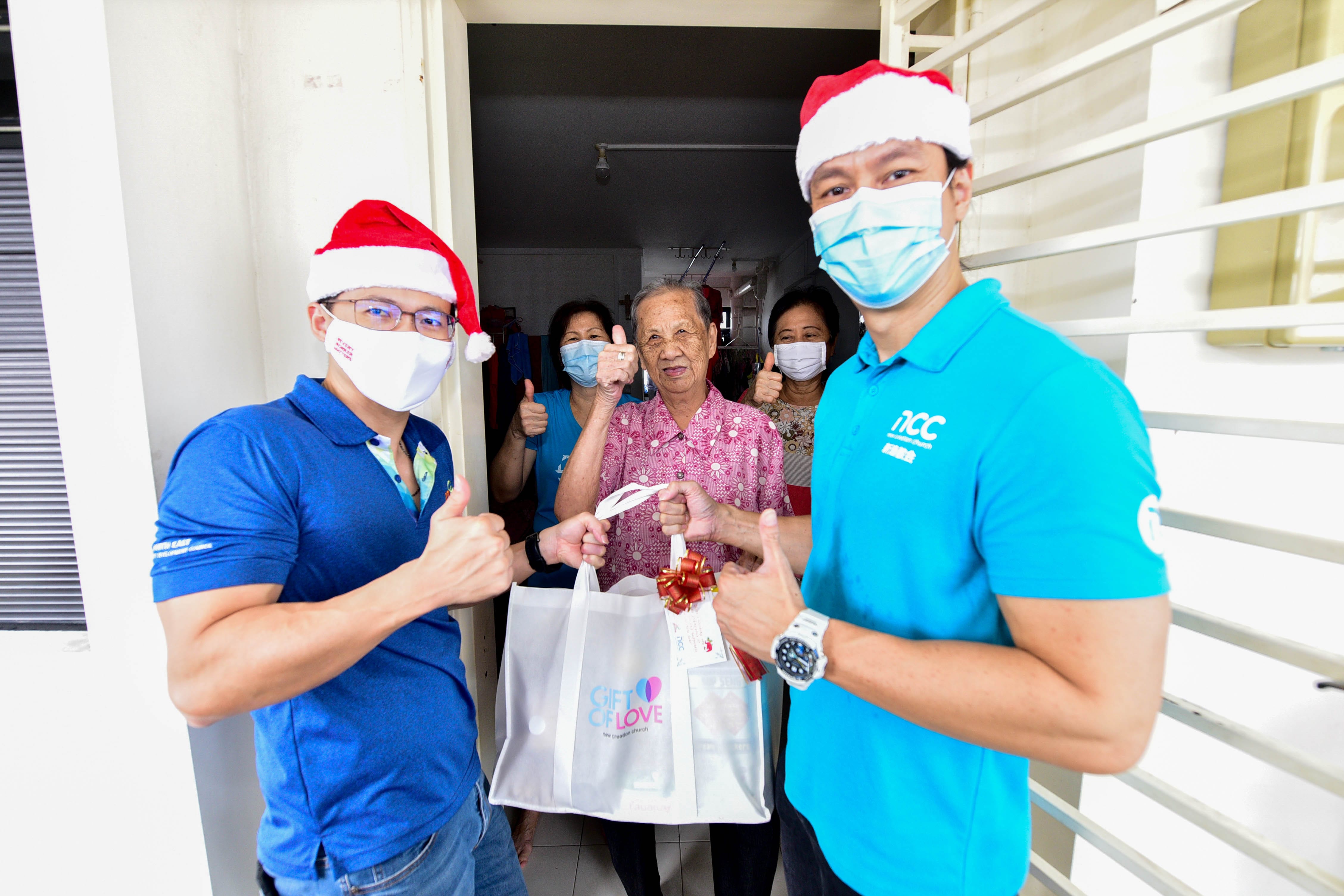 People in festive Christmas hats give a gift bag to others at a doorway, all showing thumbs up.